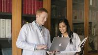 Two medical students looking at a computer and text book in a library.